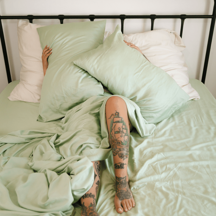 Person liegt im Bett und trägt grüne und weiße Bambus-Bettwäsche mit sichtbaren Beintattoos