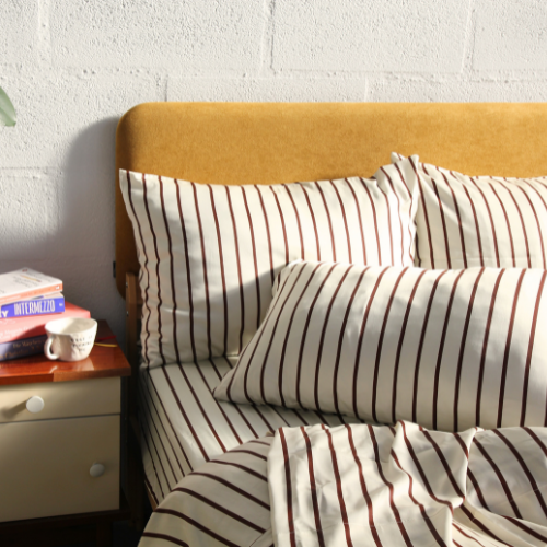 Par de fronhas de bambu acetinado às riscas, em cama com cabeceira mostarda, junto a livros e cabeceira cinzenta.
