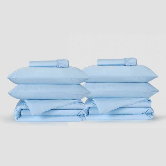 Zwei hellblaue Bettwäscheset von Momo Cotton stapelweise mit Kissen und gefalteter Bettwäsche vor neutralem Hintergrund